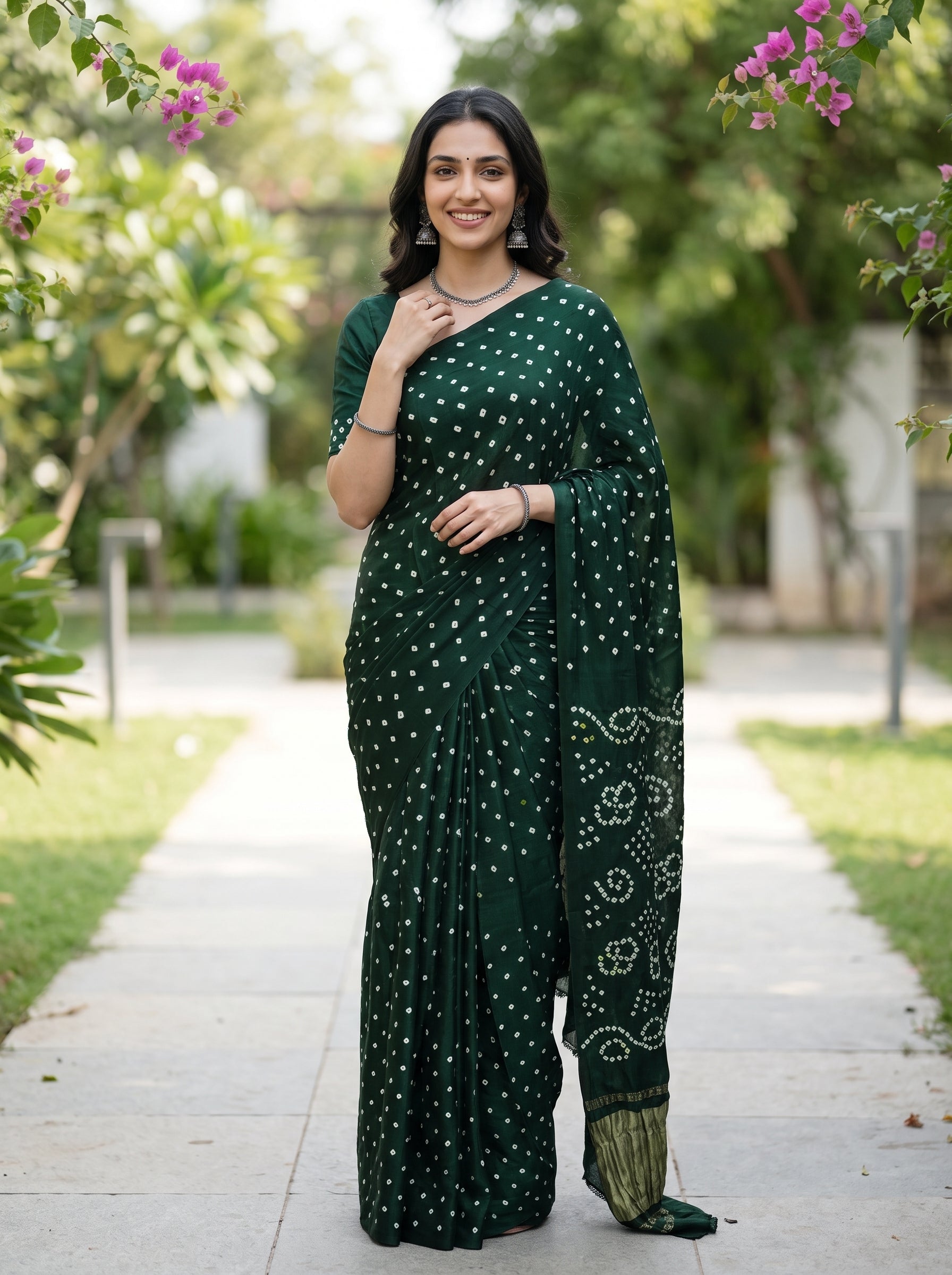 A woman wearing a dark green saree with white tie-dye bandhani patterns stands on a paved path in a garden setting.
