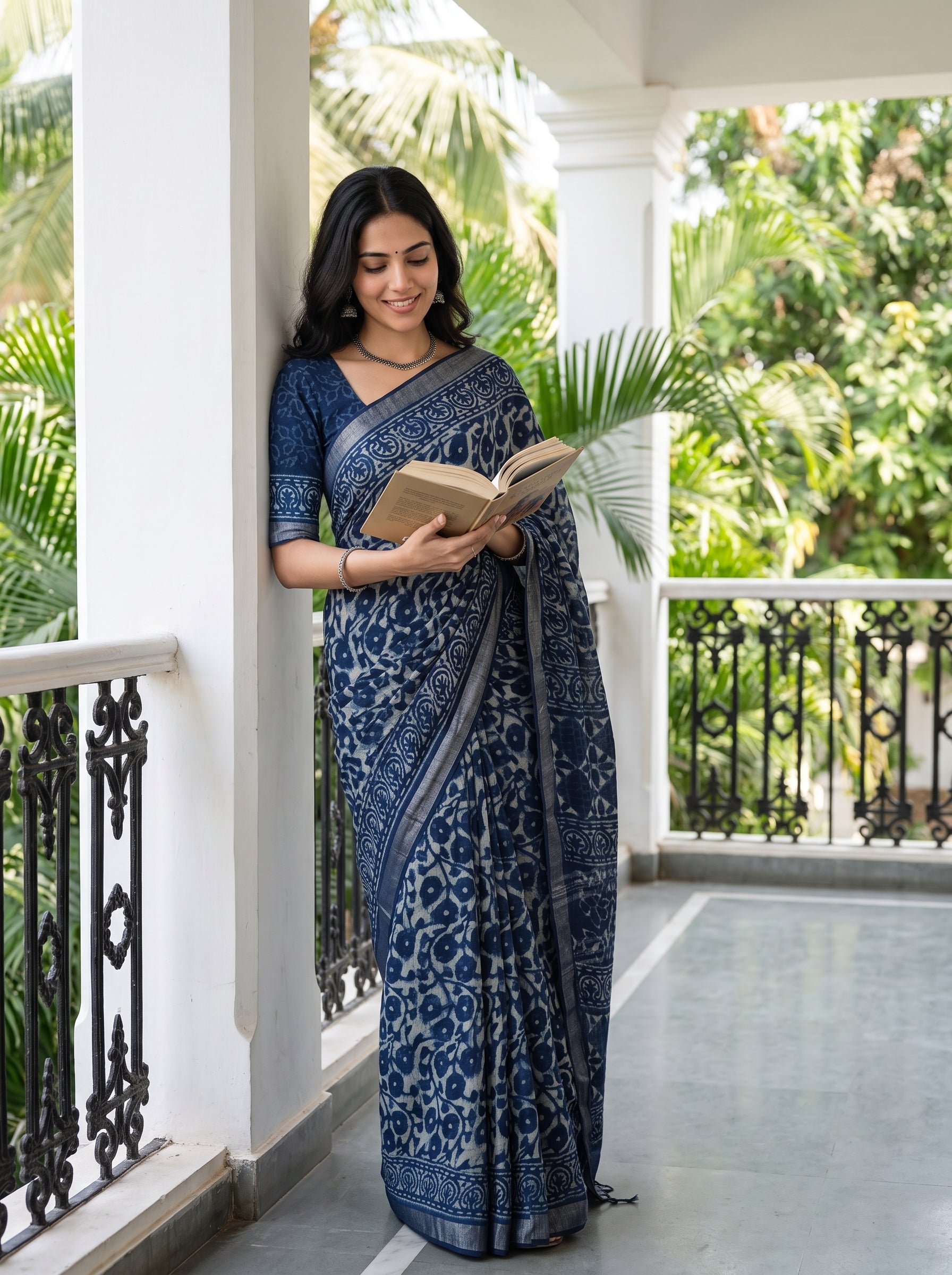 A woman wearing a navy blue and gray printed saree stands on a white-pillared balcony, holding an open book. Lush green tropical foliage is visible in the background.