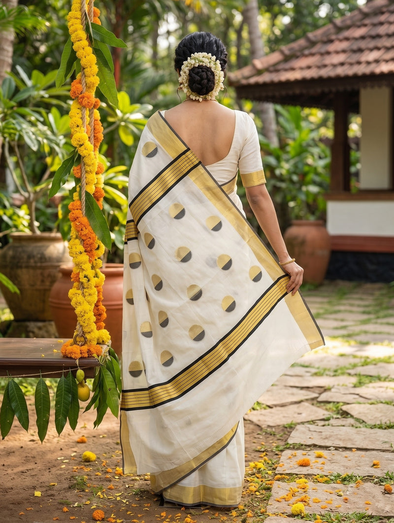 A woman wearing a white and gold saree is shown from behind in an outdoor garden setting with a stone path, potted plants, and a traditional house in the background.