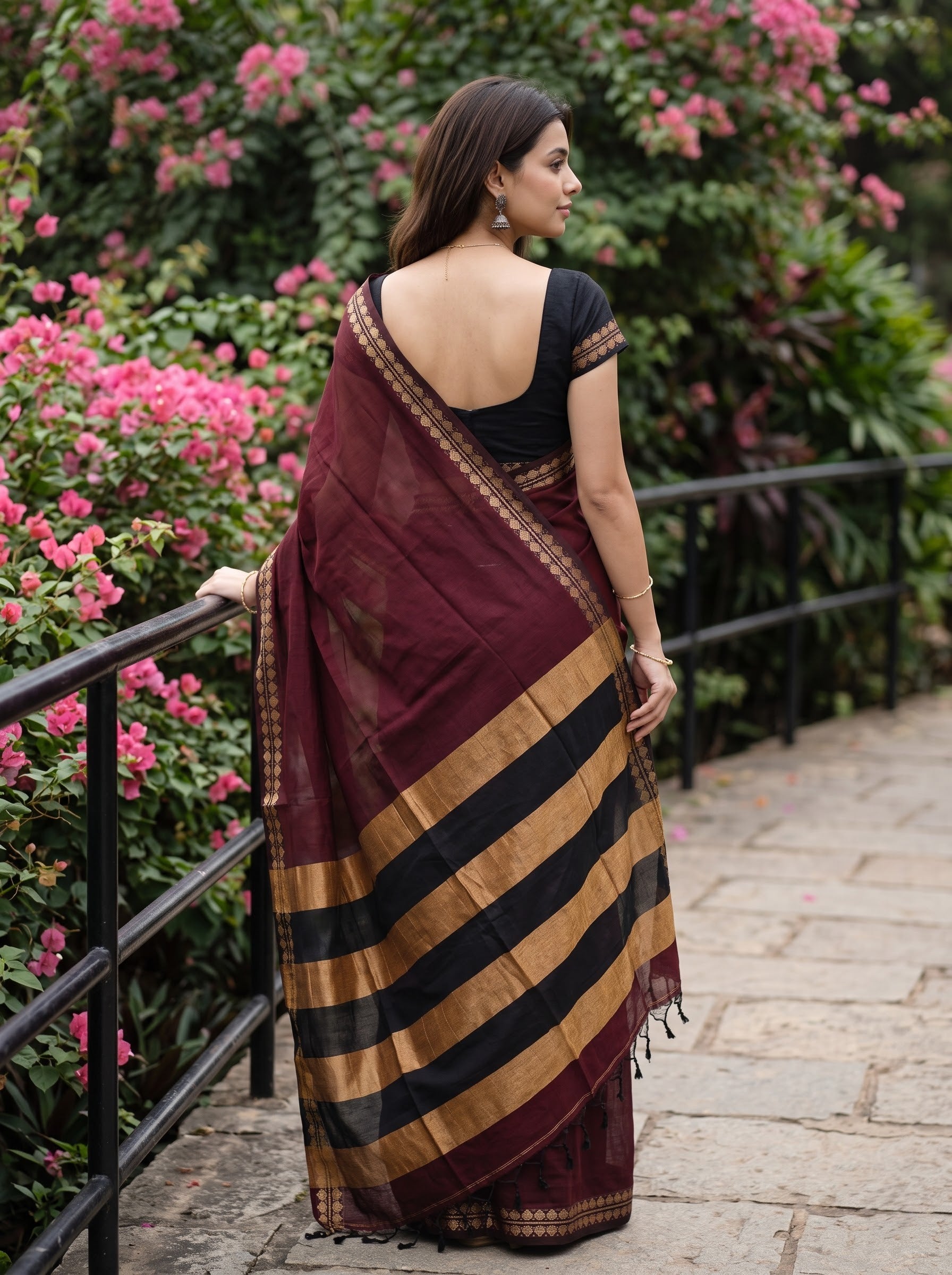 A woman wearing a deep maroon and black striped saree with gold borders, standing on a stone pathway next to a black metal railing with pink flowers in the background.