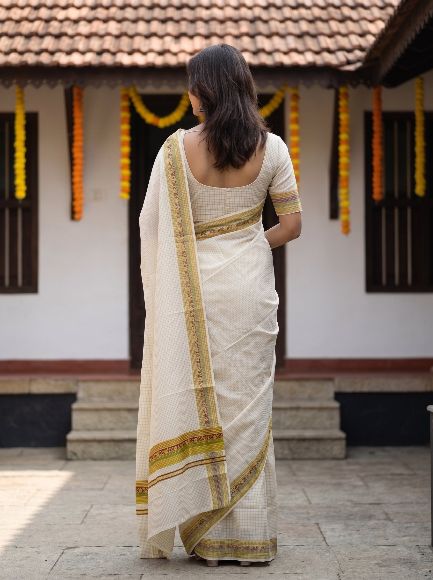 A woman viewed from behind wearing a cream-colored cotton saree with gold and green border patterns, standing in front of a traditional house with a tiled roof and marigold garlands.