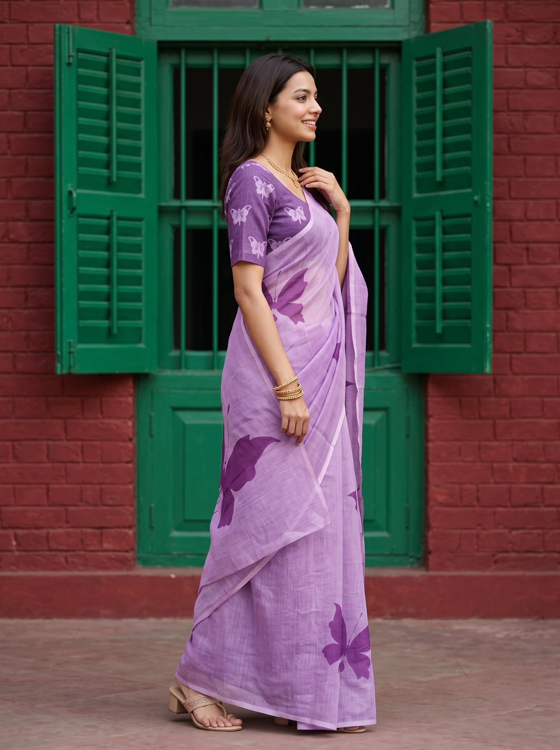 A woman wearing a lavender purple saree with butterfly prints and a matching printed blouse, standing in front of a red brick wall with green shutters.