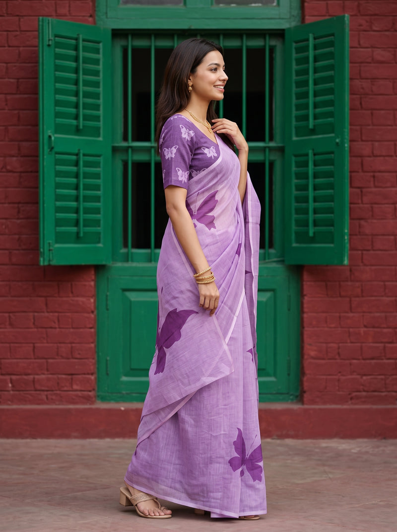 A woman wearing a lavender purple saree with butterfly prints and a matching printed blouse, standing in front of a red brick wall with green shutters.