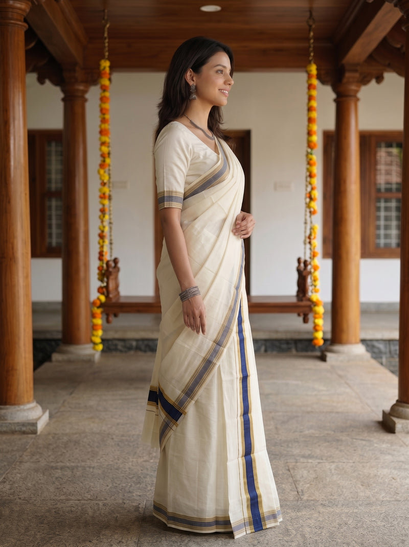 A woman wearing a cream-colored cotton saree with blue and gold stripes, standing in profile in a traditional courtyard with wooden pillars and hanging marigold garlands.