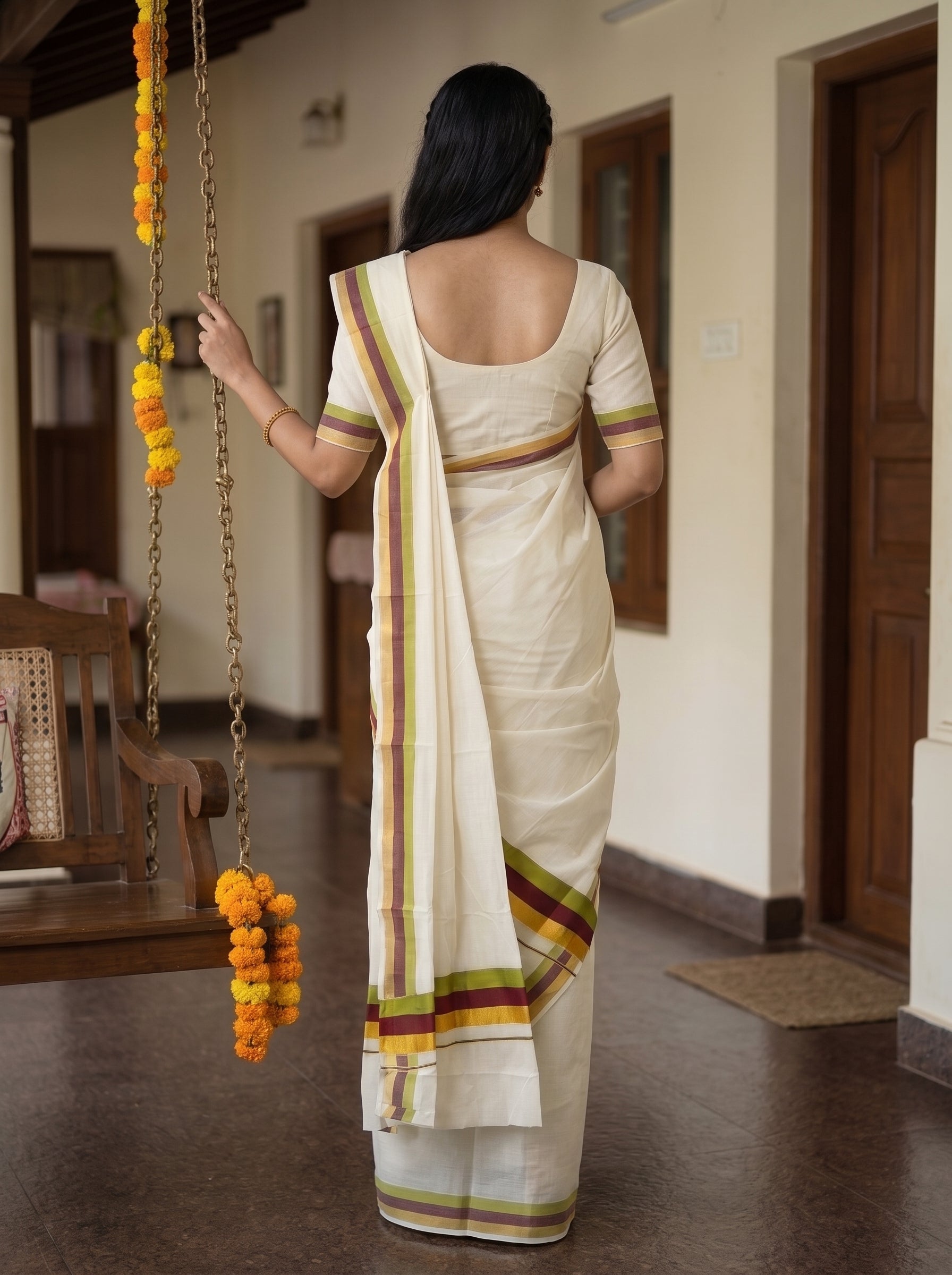A woman viewed from behind wearing a cream-colored cotton saree with a matching short-sleeve blouse. The saree features thin stripes of green, yellow, and red along the border and pallu.