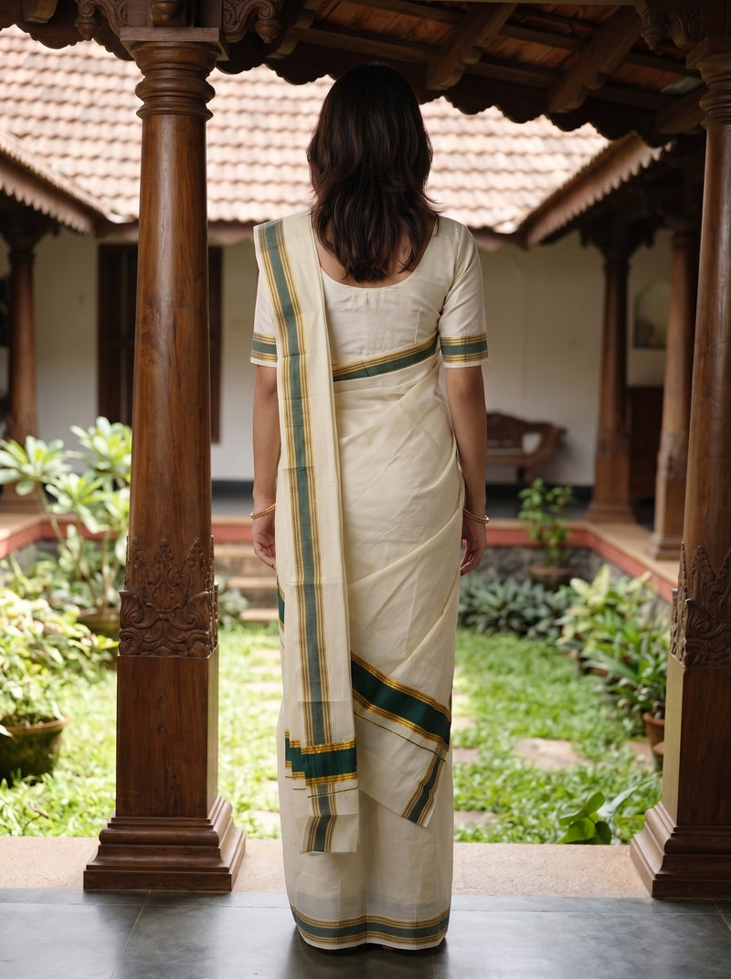 A woman viewed from behind wearing a cream-colored cotton saree with green and gold stripes, standing in a traditional courtyard framed by carved wooden pillars.
