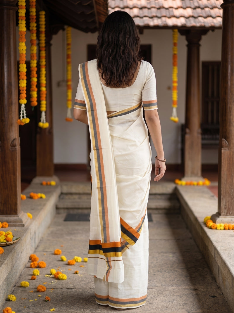 A woman viewed from behind wearing a cream-colored cotton saree with orange, yellow, and black striped borders, standing in a traditional courtyard decorated with marigold garlands.