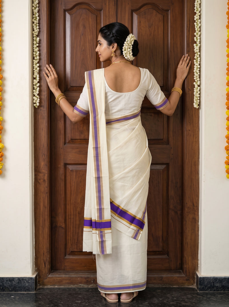 A woman wearing a cream-colored cotton saree with purple and gold borders, standing in front of a dark wooden door and touching the door with both hands.