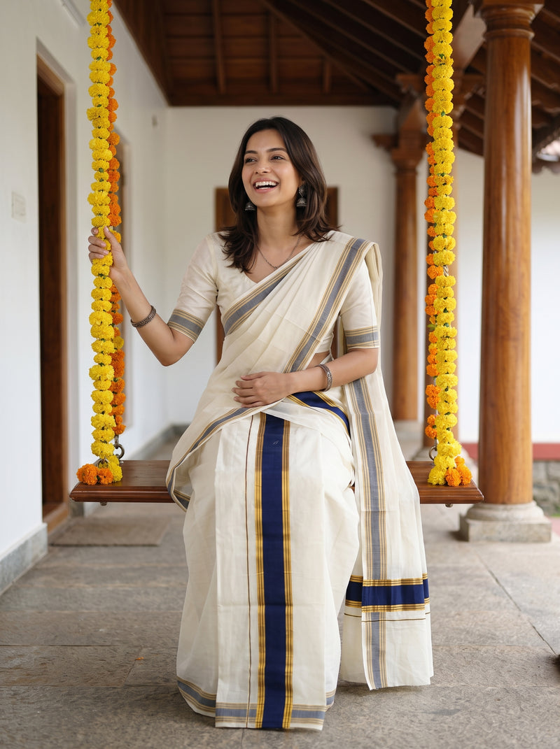 A woman wearing a cream-colored cotton saree with navy blue and gold stripes, standing on a traditional wooden swing decorated with yellow flower garlands.