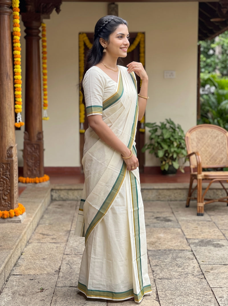 A woman wearing a cream-colored cotton saree with green and gold borders, standing in a traditional courtyard setting.