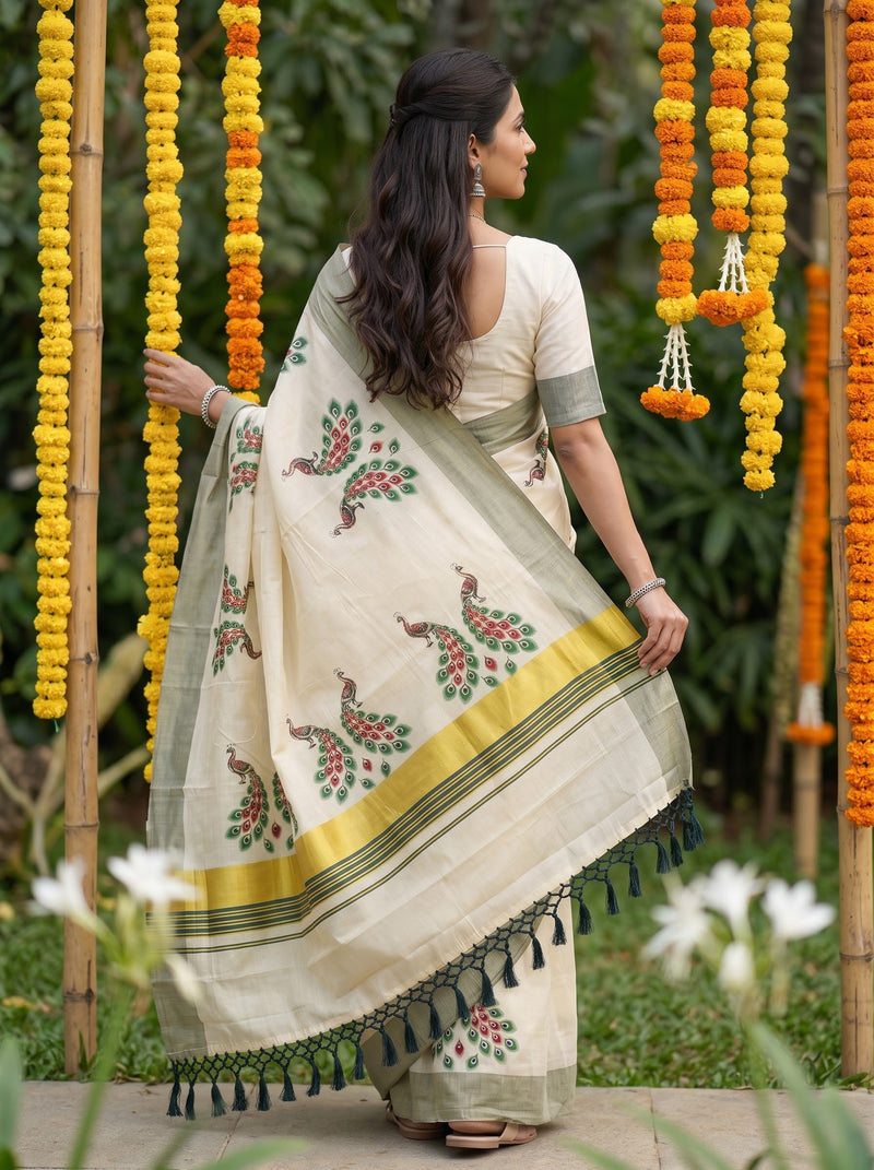 A woman wearing a cream-colored saree with green and red floral motifs and a yellow border, standing outdoors in front of bamboo garlands.
