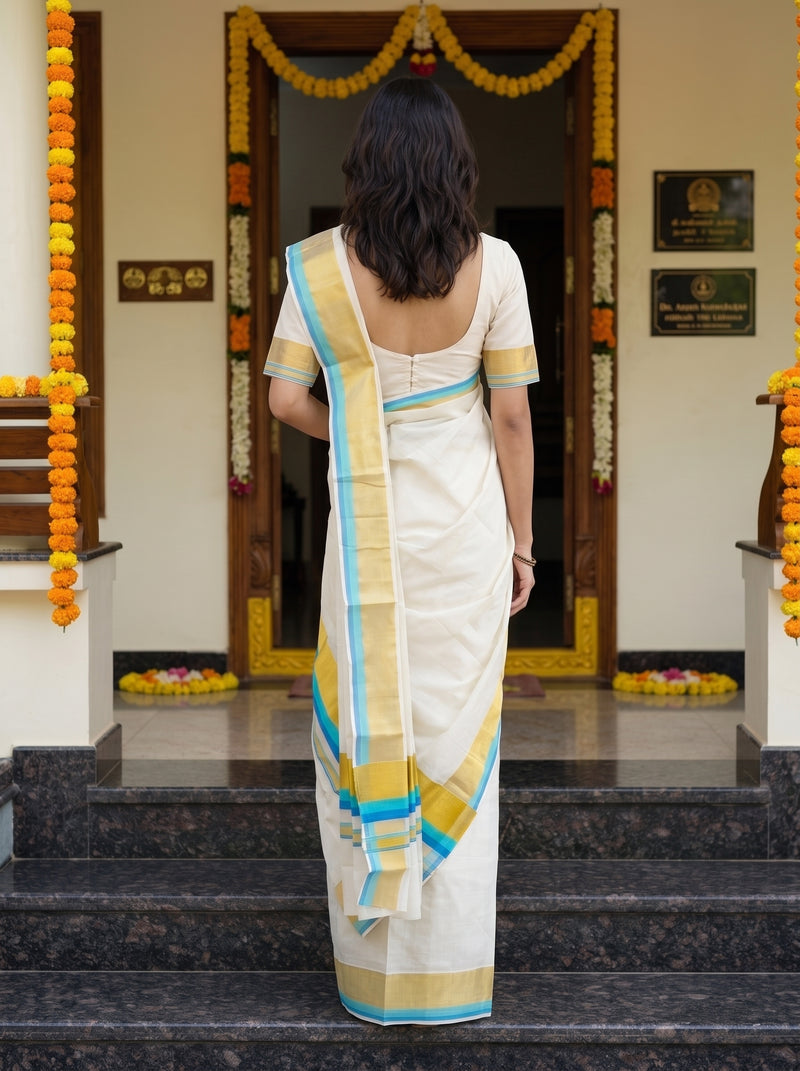 A woman viewed from behind wearing a white saree with a yellow and blue border, standing on dark stone steps in front of a decorated doorway with marigold garlands.
