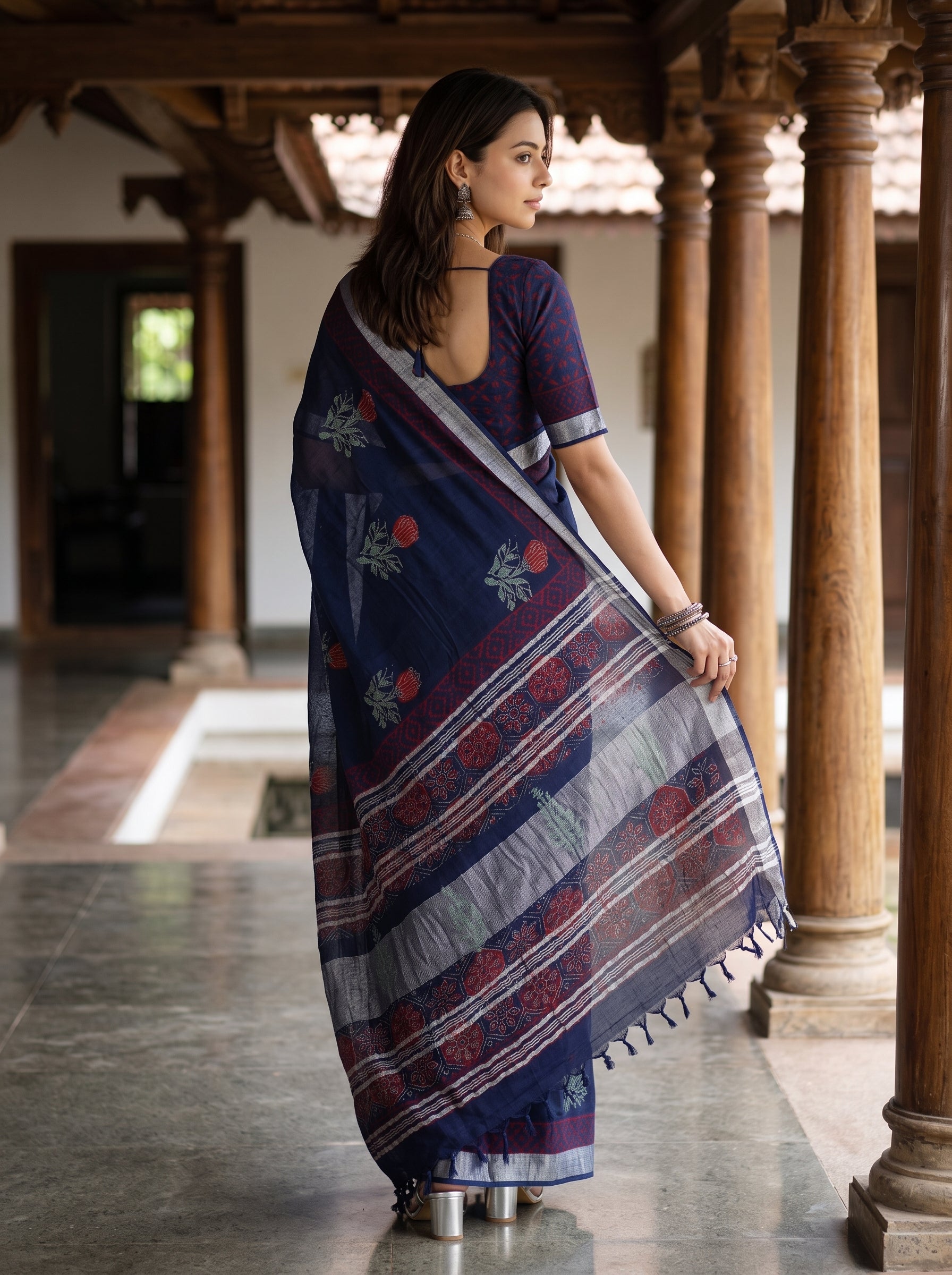 A woman wearing a navy blue and purple printed saree with a matching purple blouse, standing in a traditional courtyard with wooden pillars.