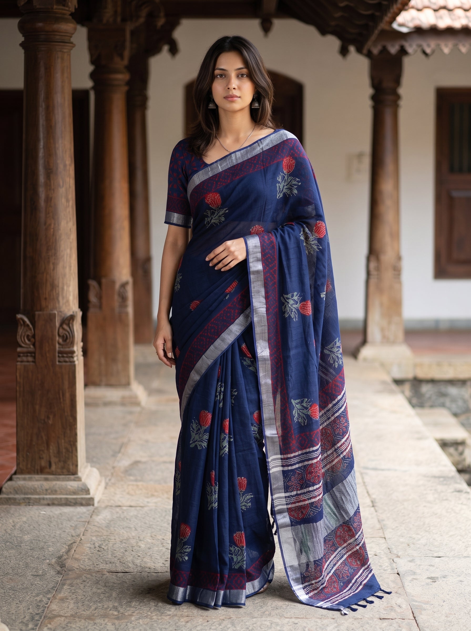 A woman wearing a navy blue printed saree with red and silver floral motifs, standing in a traditional courtyard with wooden pillars.