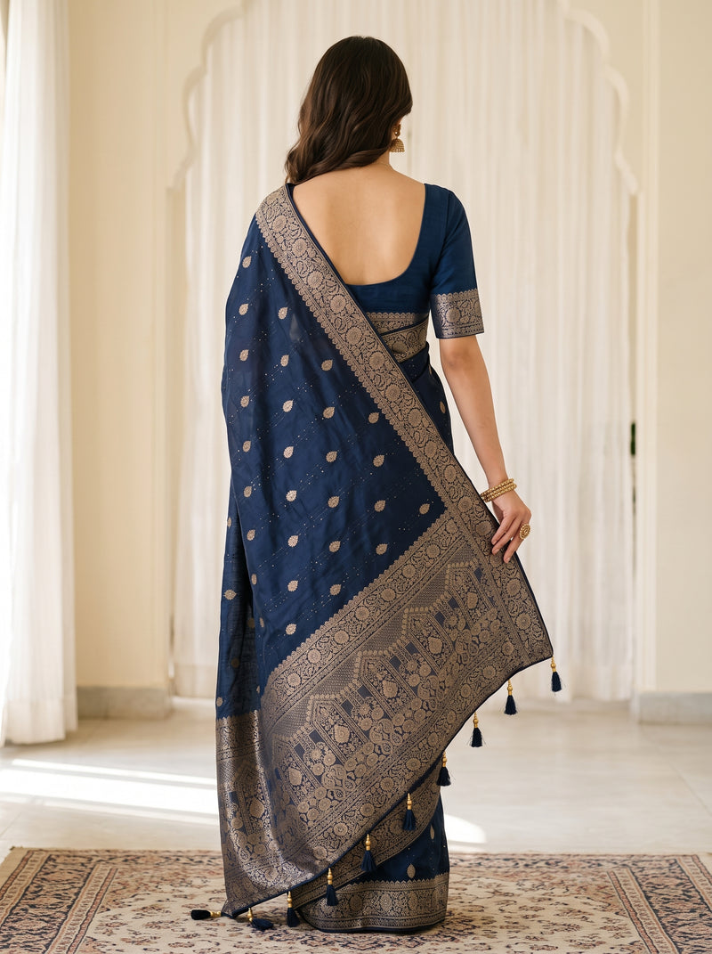 A woman viewed from behind wearing a navy blue saree with gold zari motifs and a wide ornate border, standing indoors on a patterned rug in front of cream-colored arched doorways.