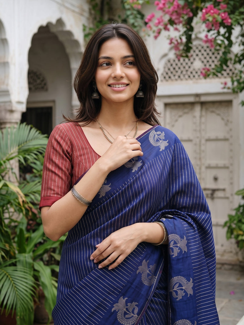 A woman wearing a royal blue saree with silver zari motifs and a matching maroon blouse, standing in an outdoor courtyard with architectural details and greenery.