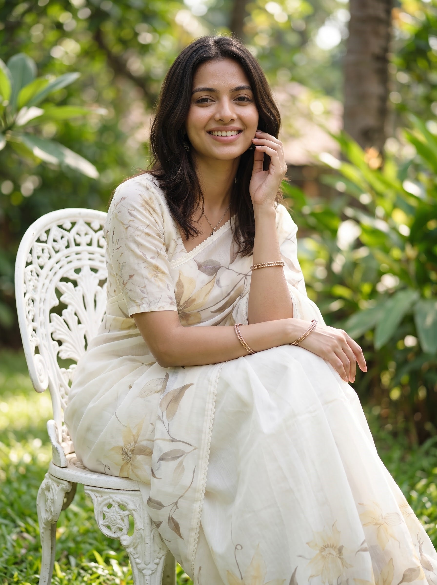 A woman wearing a cream-colored floral printed saree sits on a white garden chair outdoors, surrounded by green foliage.