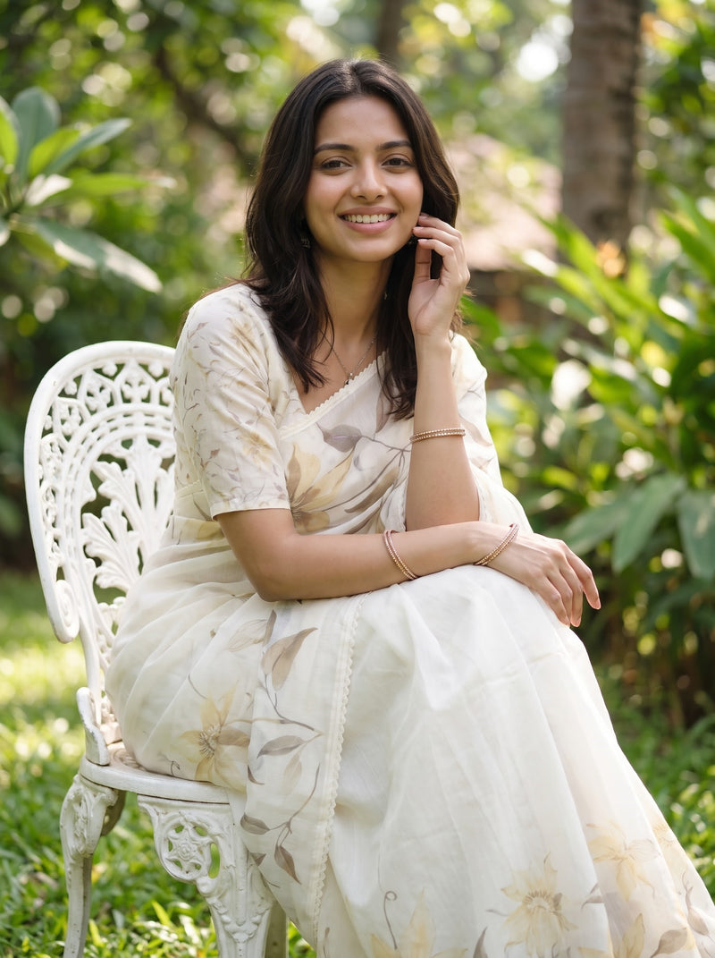 A woman wearing a cream-colored floral printed saree sits on a white garden chair outdoors, surrounded by green foliage.