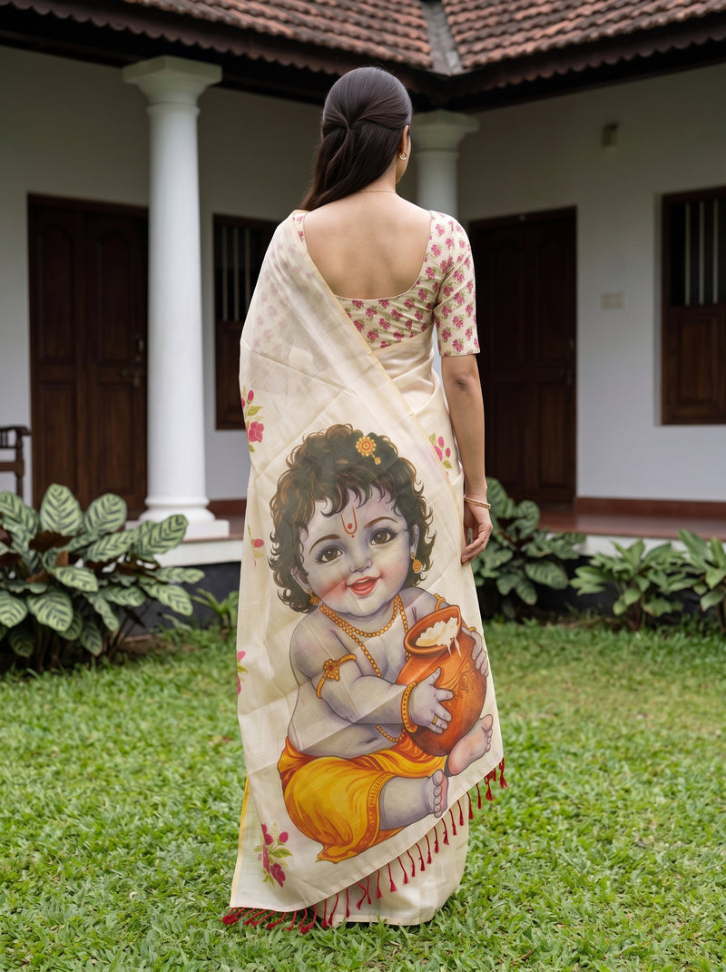 A woman stands outdoors in front of a house, shown from the back wearing a cream-colored saree with a large printed illustration of a child holding an orange pot. The saree has red and yellow accents, and she is paired with a patterned blouse.