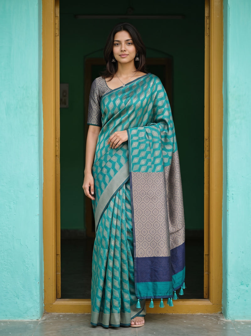 A woman stands in a doorway wearing a teal and gray patterned saree with a dark gray blouse. The background features a turquoise wall and a dark interior.