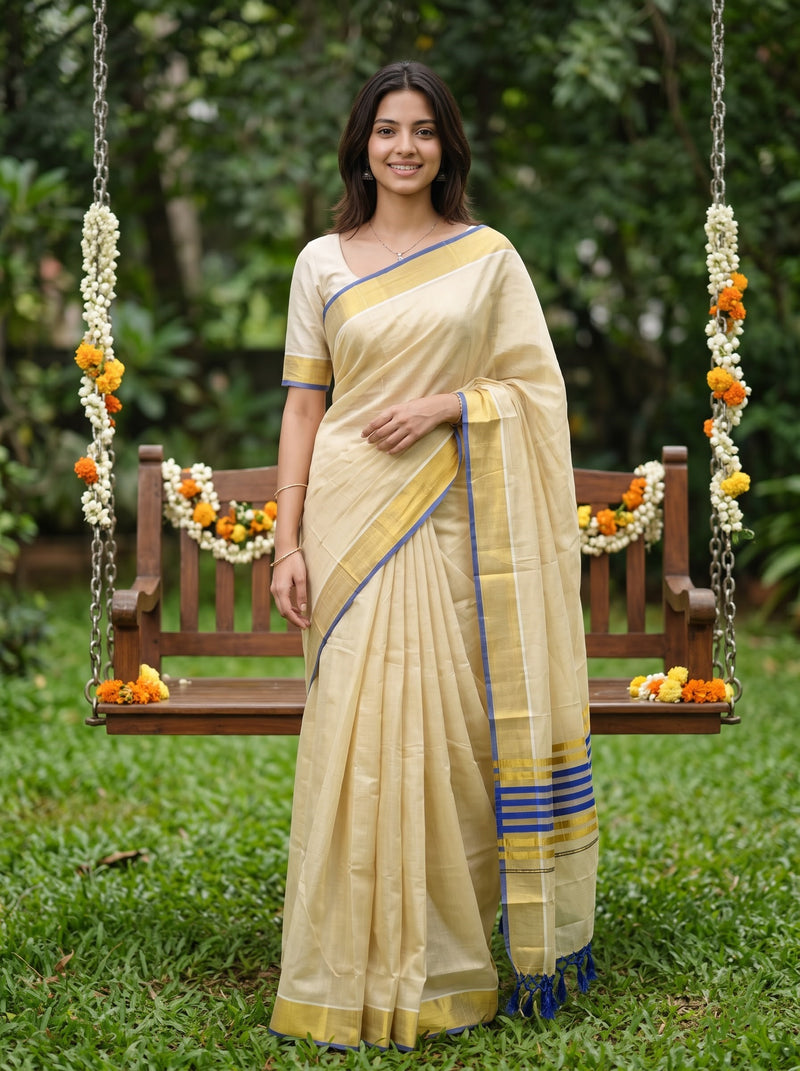 A woman wearing a cream-colored silk saree with blue and gold borders stands on a wooden swing decorated with marigold and white flower garlands in a lush green garden setting.