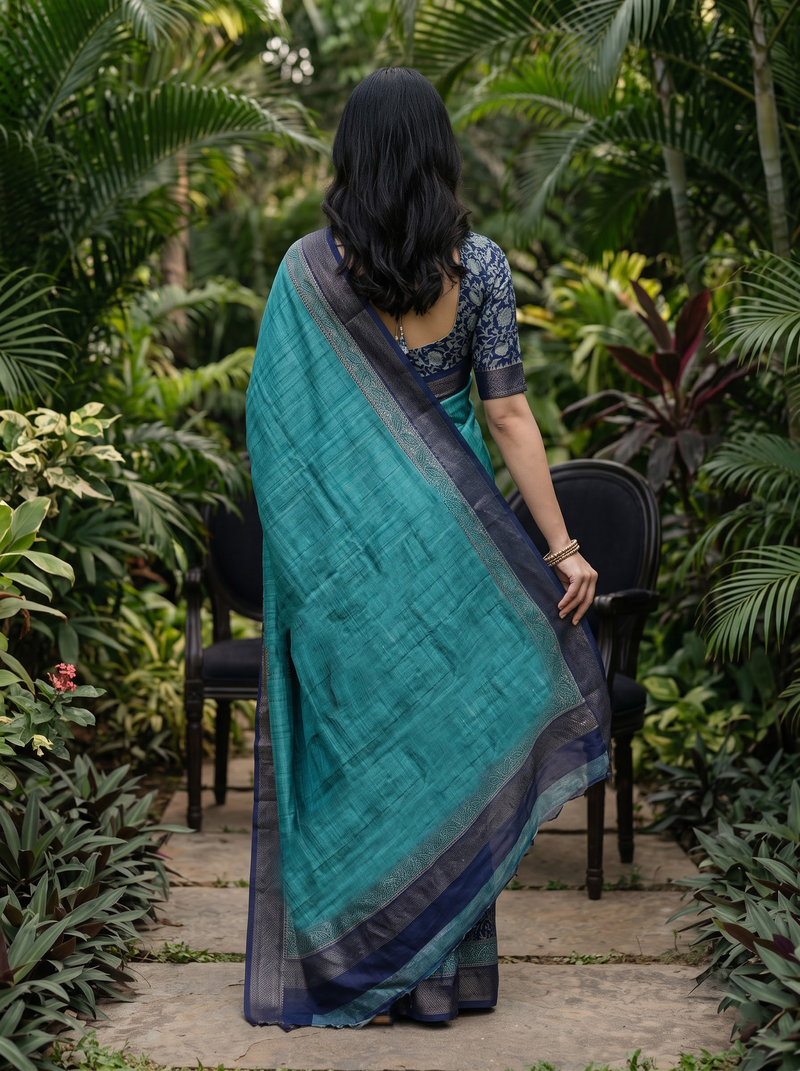 A woman stands with her back to the camera in a lush garden setting, wearing a teal and navy blue saree with a patterned blouse.