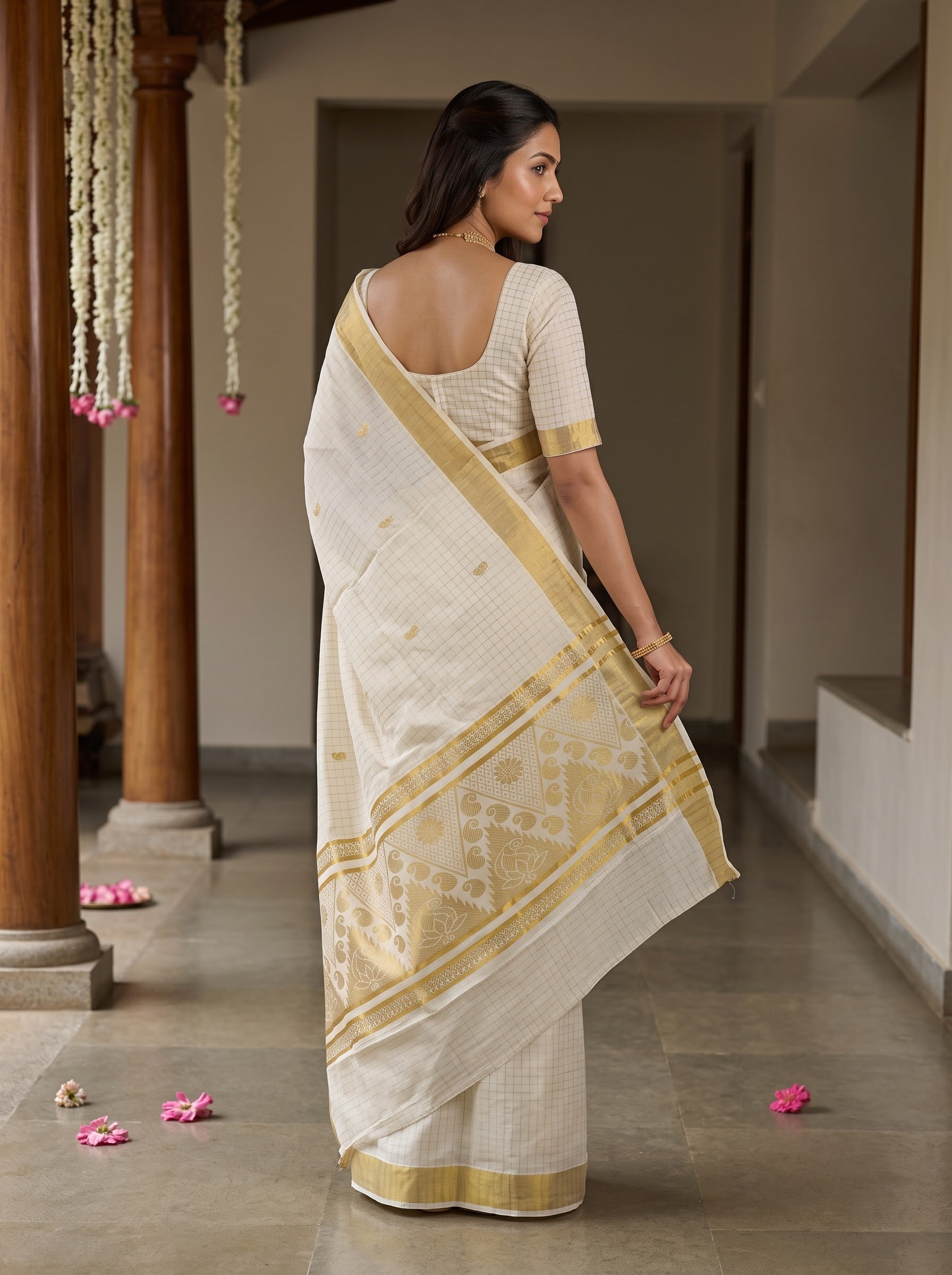A woman wearing a cream-colored cotton saree with a gold border and matching cream blouse, standing in a corridor with wooden pillars.