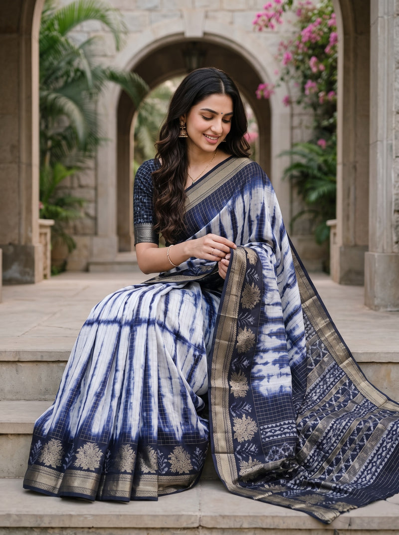A woman seated on stone steps wearing a navy and white tie-dye saree with a patterned blouse, photographed in an outdoor architectural setting with arches and plants.
