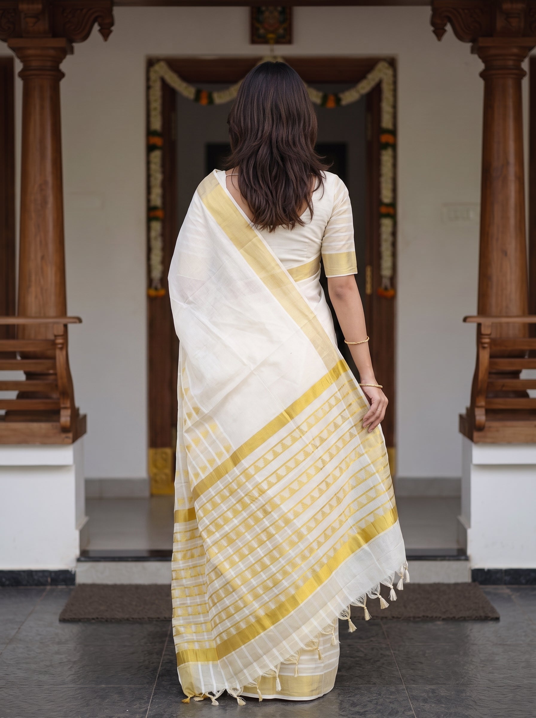 A woman viewed from behind wearing a white and yellow checkered saree with a yellow border, standing in front of a traditional doorway with wooden pillars.