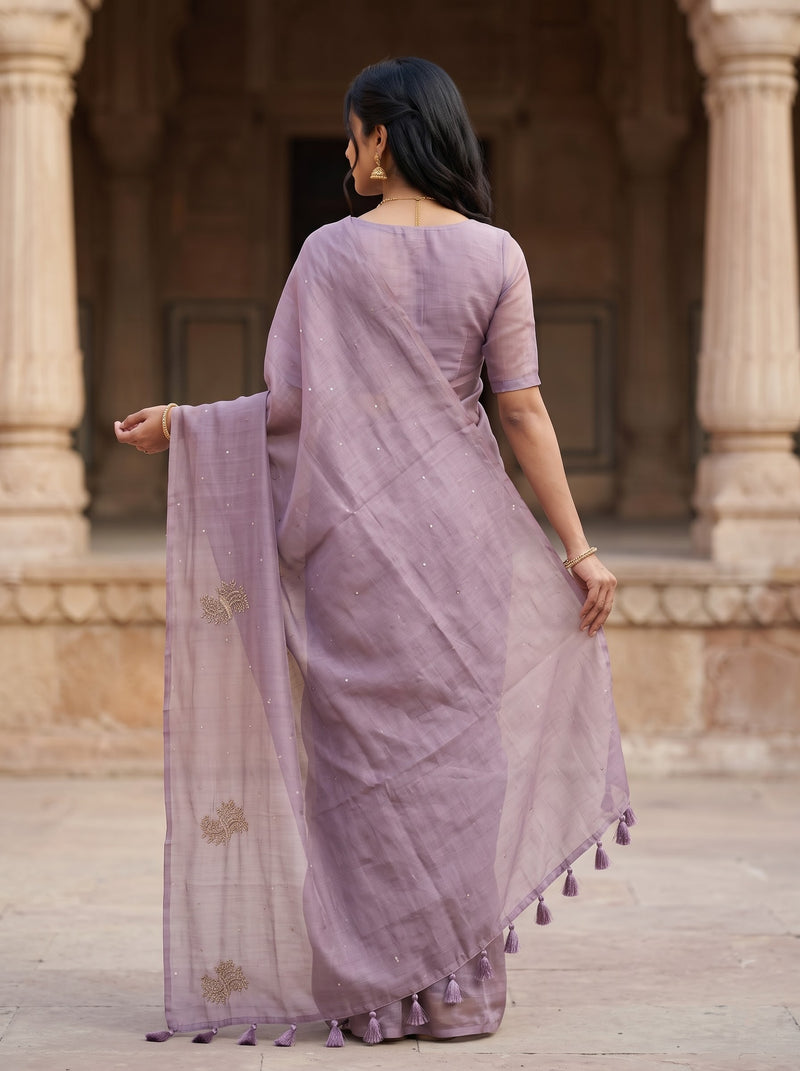 A woman wearing a lavender purple saree with a matching blouse, shown from behind in front of a stone architectural background with pillars.