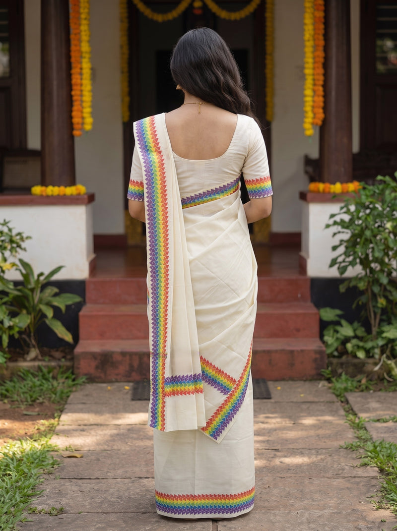 A woman viewed from behind wearing a cream-colored saree with colorful rainbow-patterned borders, standing in front of a decorated doorway with steps and plants.