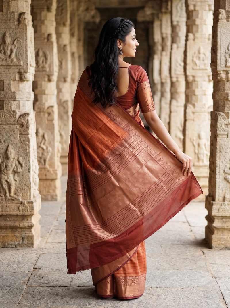 A woman wearing a rust-red and copper-brown silk saree with a matching blouse, standing in a stone corridor framed by carved pillars.