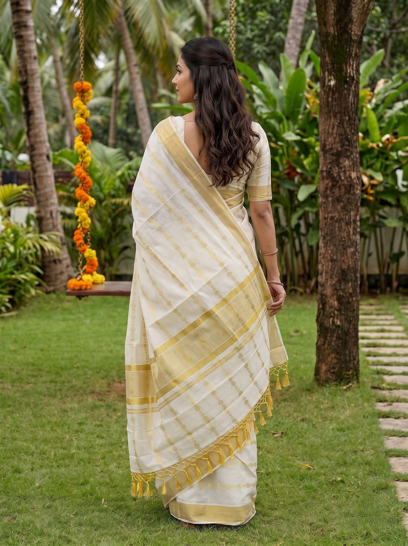A woman wearing a cream and gold saree stands outdoors in a garden, shown from the back. The saree has a cream base with gold stripes and a gold border, and the background features green grass, trees, and a hanging swing decorated with orange marigold flowers.