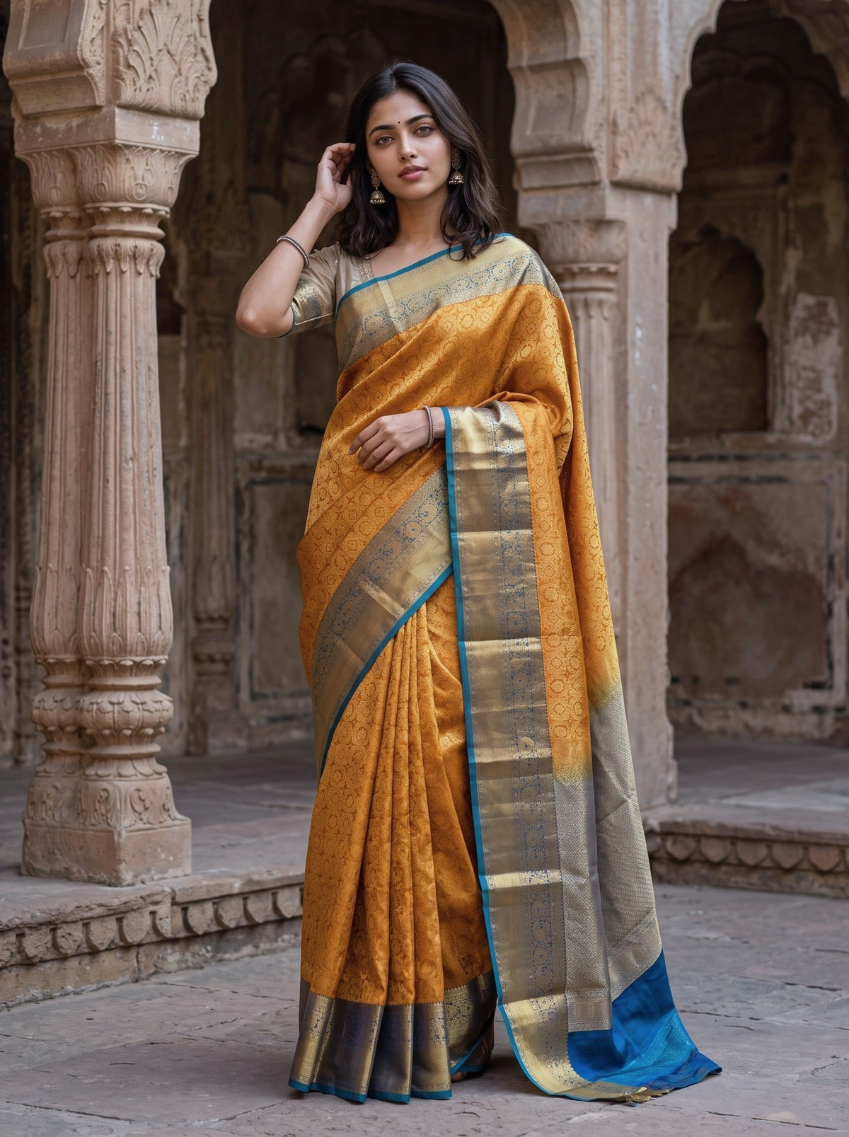 A woman models a golden yellow silk saree with a blue border and a silver zari border, standing in front of a historic stone archway.