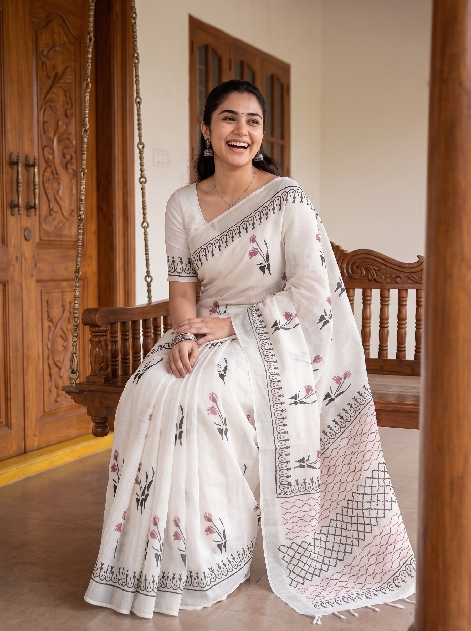 A woman wearing a white embroidered saree with pink floral motifs and black geometric border patterns, seated on a wooden swing in a warm indoor setting.
