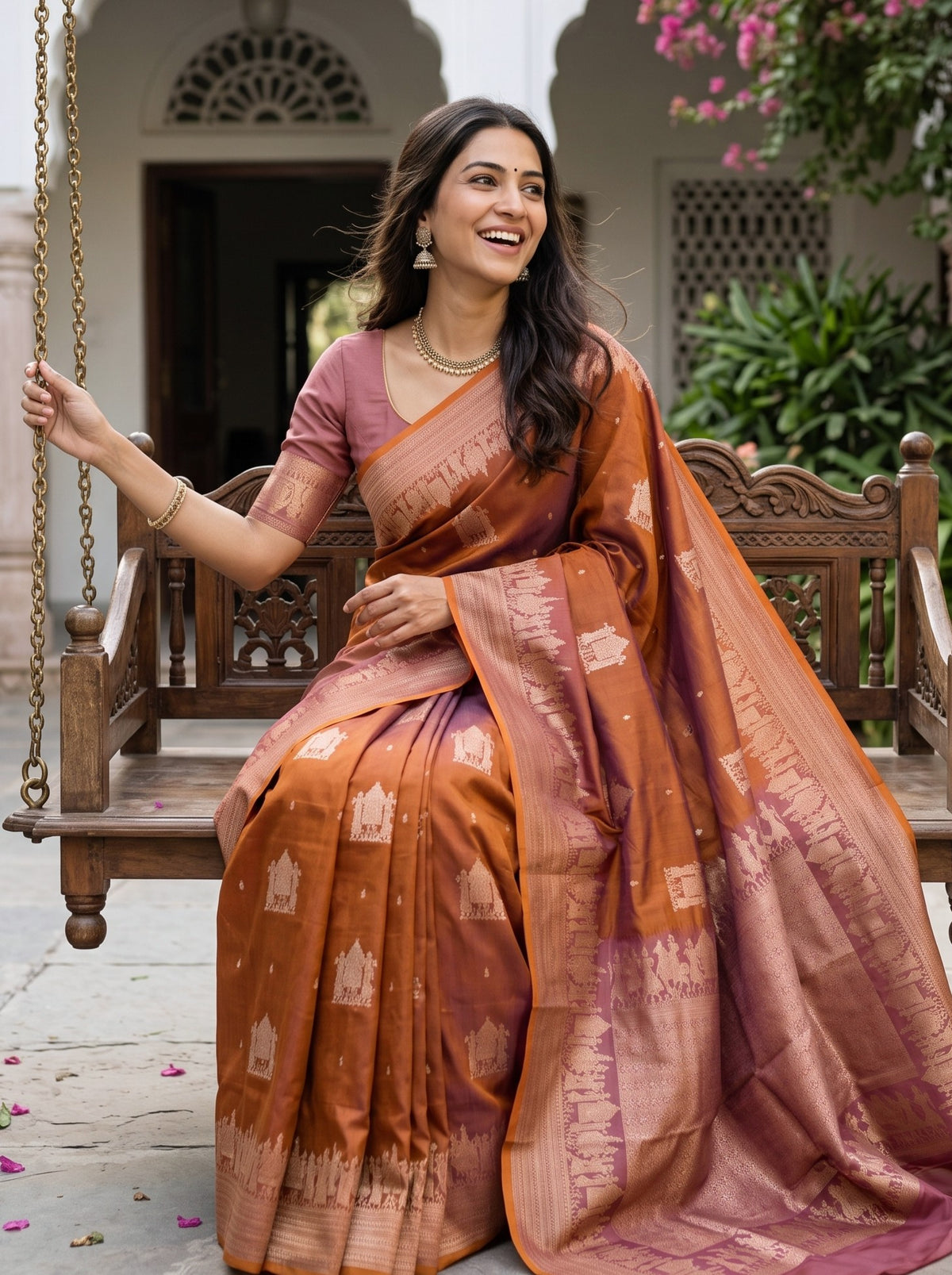 A woman wearing a rust-orange and mauve Banarasi silk saree with white zari motifs, sitting on a wooden swing bench in an outdoor courtyard.
