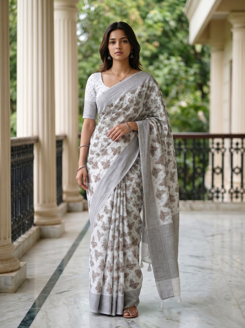 A woman models a light gray and white saree with brown floral patterns, paired with a matching white blouse, standing on a marble walkway with columns and green foliage in the background.