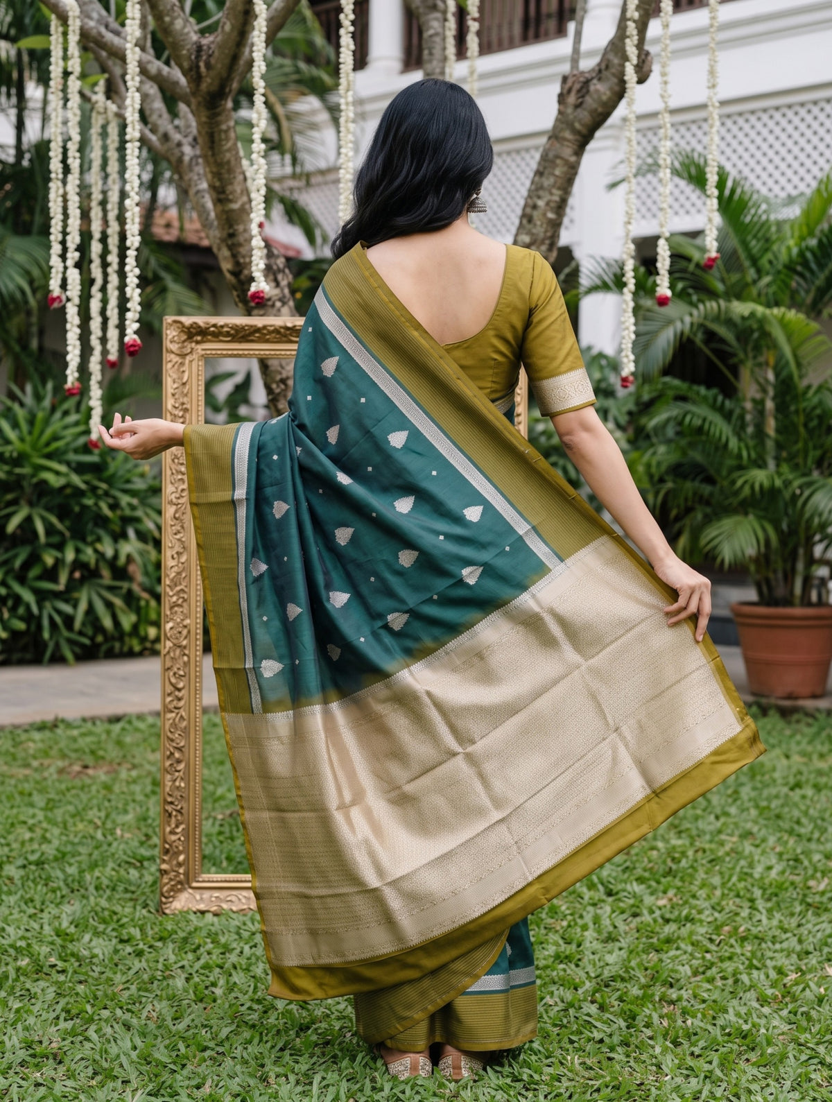 A woman stands outdoors on grass holding up a green and beige saree to show its drape and pallu. The saree has a deep green body with small white motifs and a wide beige border, paired with a matching olive green blouse.