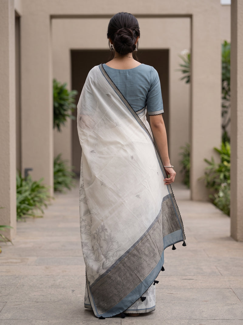 A woman viewed from behind wearing a white and gray saree with a dusty blue short-sleeve blouse, standing in a stone walkway with plants.