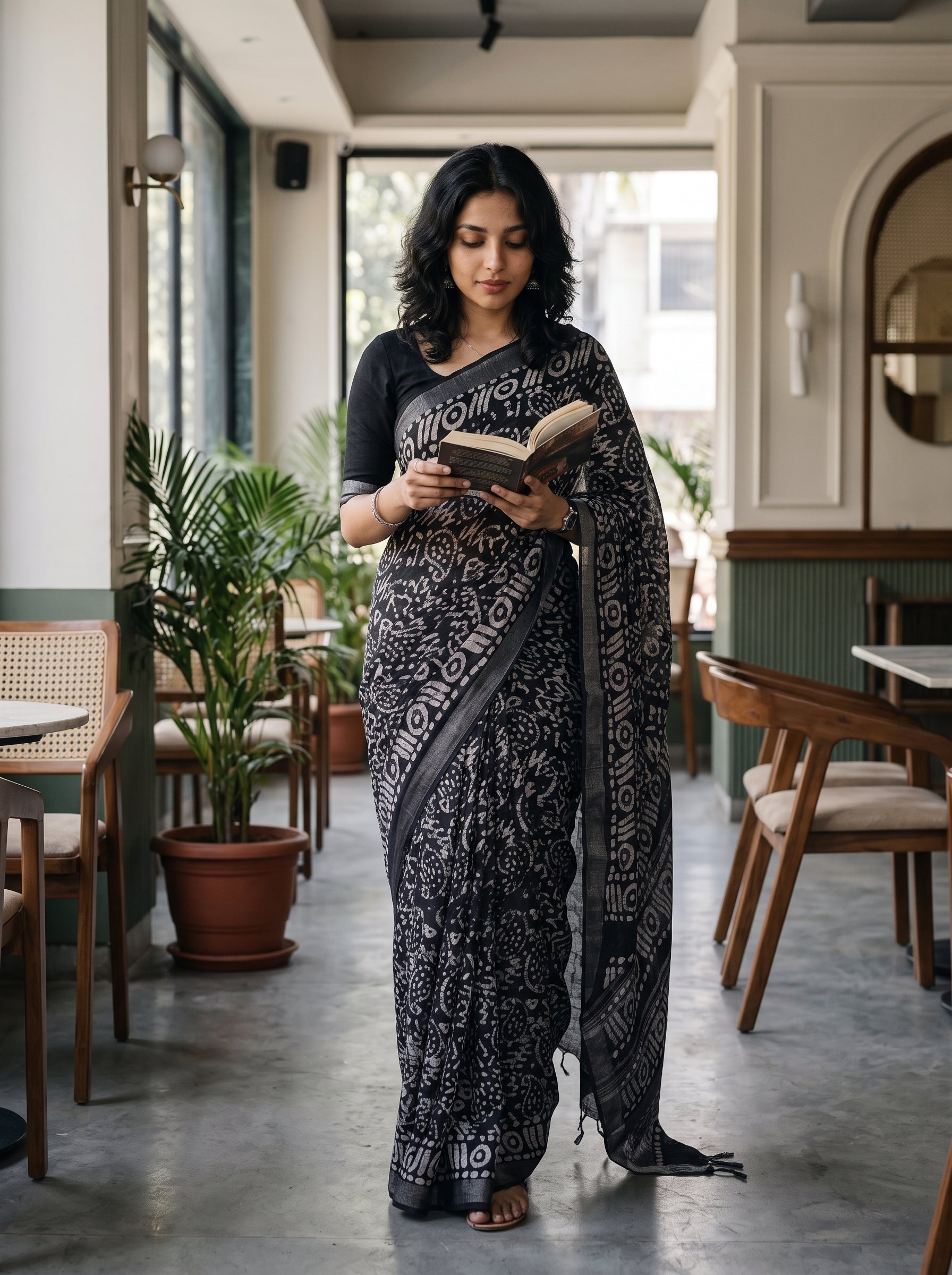 A woman wearing a black and gray patterned sari stands in a cafe reading a book. The sari features a black base with intricate gray and white floral motifs, paired with a solid black blouse.