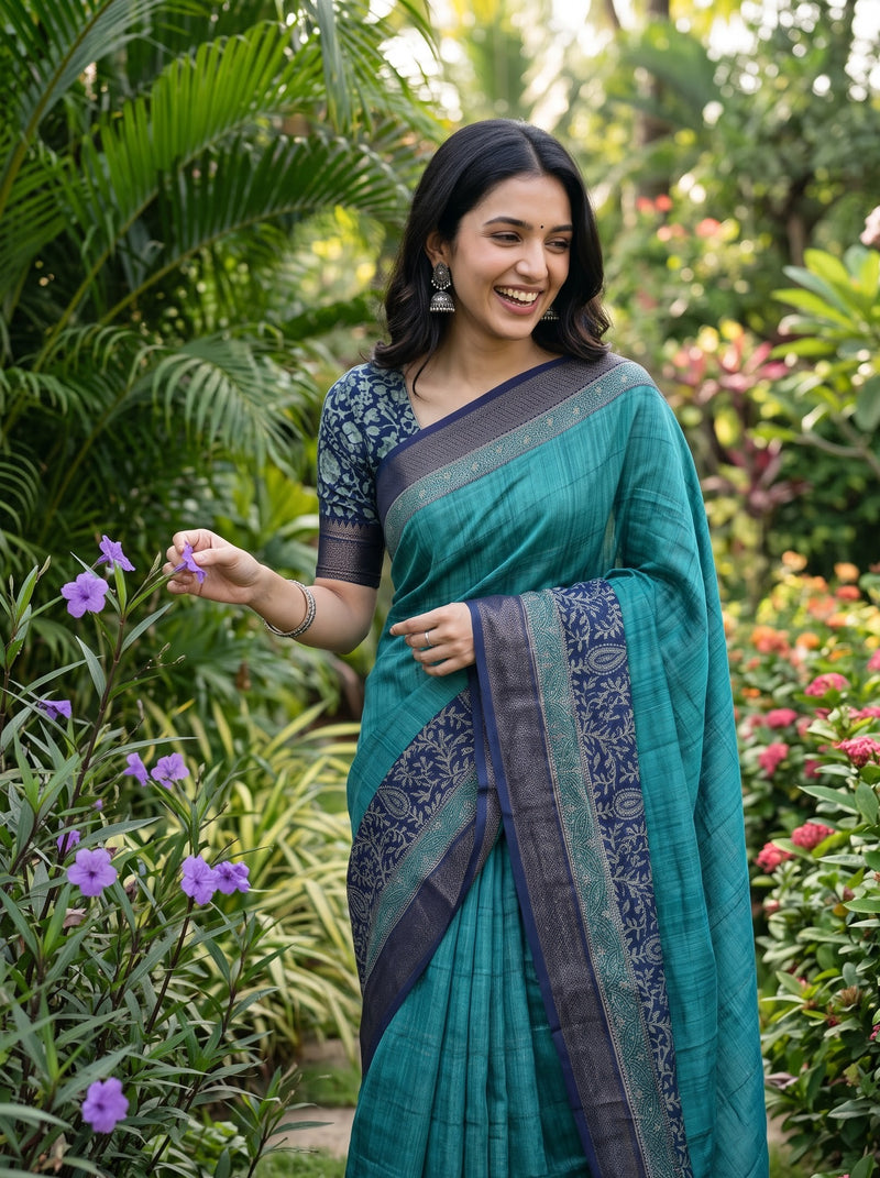 A woman wearing a teal green and navy blue saree with a patterned blouse stands in a lush garden, smiling and reaching toward small purple flowers.