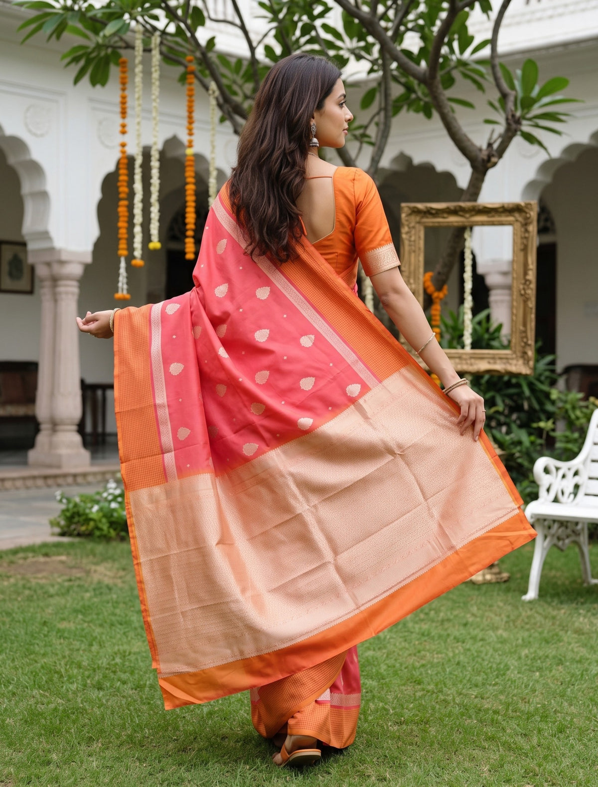 A woman wearing a peach and orange saree with gold motifs, standing in a garden courtyard and holding out the fabric to display its drape and border.
