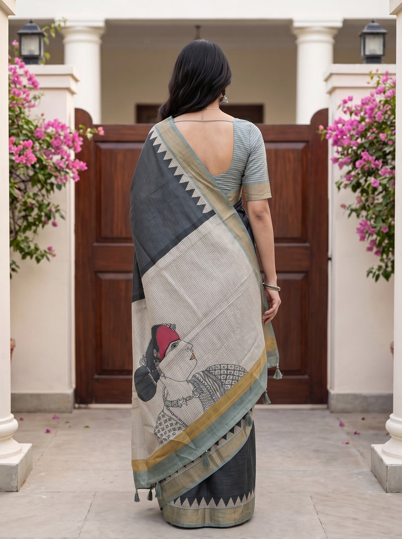 A woman stands with her back to the camera wearing a black and beige saree with a printed illustration of a woman in a red hat, set against a courtyard backdrop with wooden doors and pink flowers.