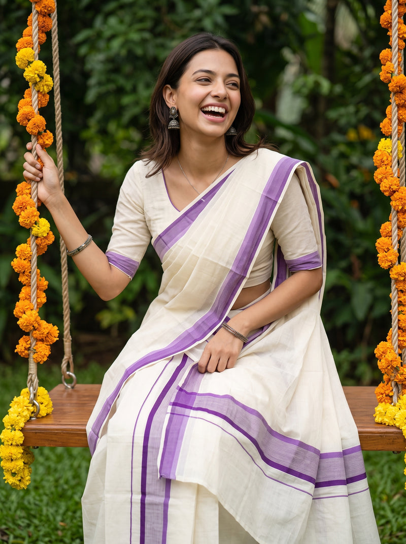 A woman wearing a cream-colored cotton saree with purple borders sits on a wooden swing decorated with orange marigold garlands, set against a lush green garden background.