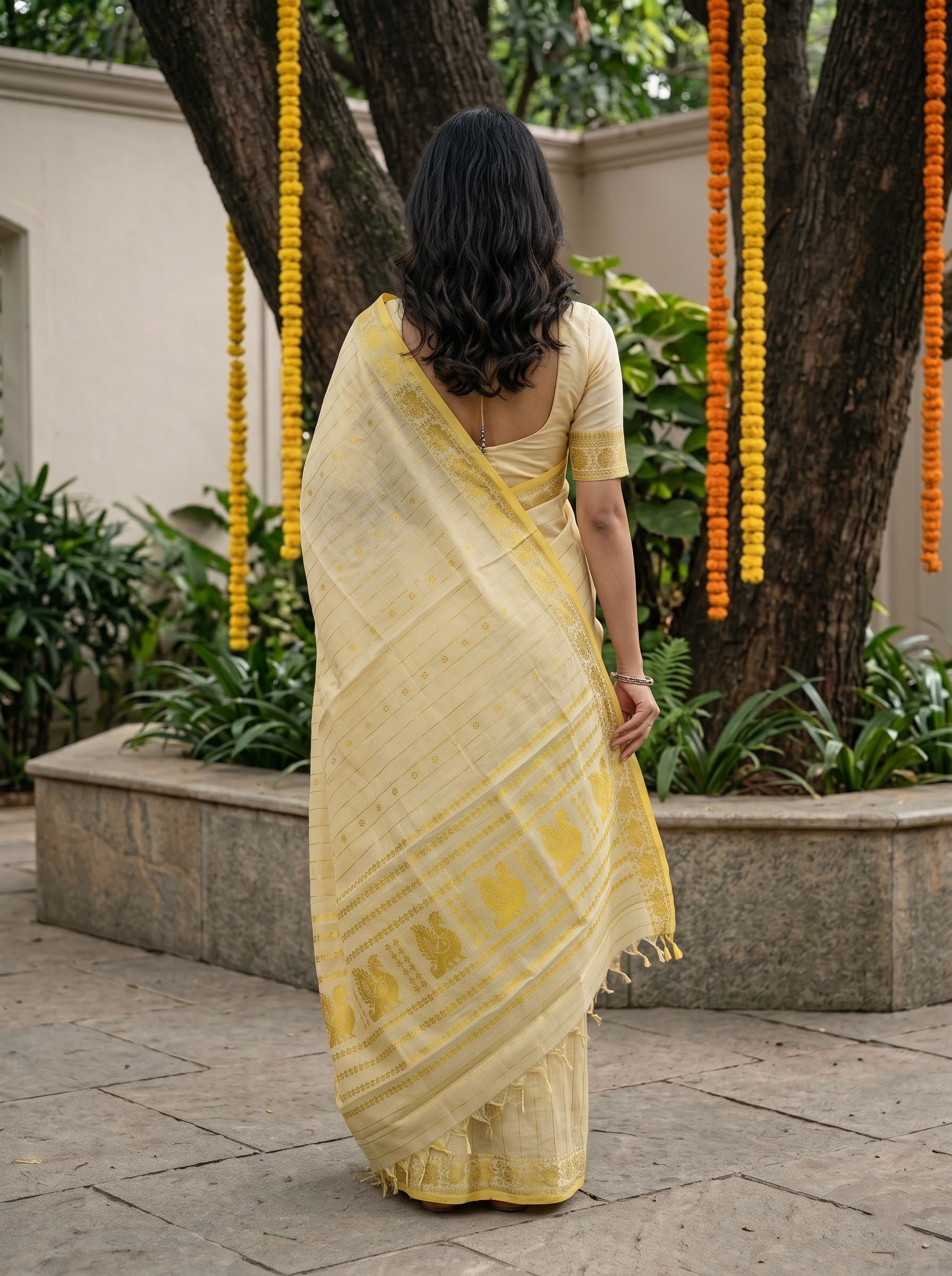A woman viewed from behind wearing a pale yellow saree with a matching blouse, standing outdoors in front of a tree decorated with yellow and orange marigold garlands.