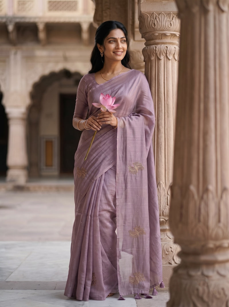 A woman wearing a lavender-purple saree with gold floral embroidery stands in a historic stone courtyard, holding a pink lotus flower.