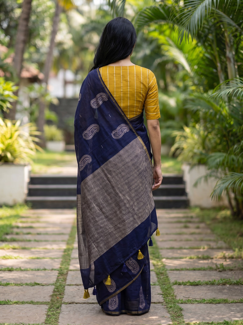 A woman viewed from behind wearing a navy blue and gray patterned saree with a mustard yellow striped short-sleeve blouse, standing on a stone path in a lush green garden.