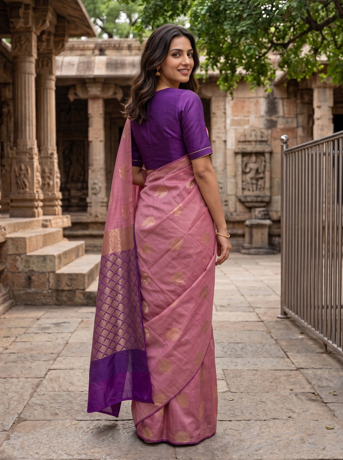 A woman wearing a pink and purple saree with gold motifs, standing in front of a stone temple courtyard.