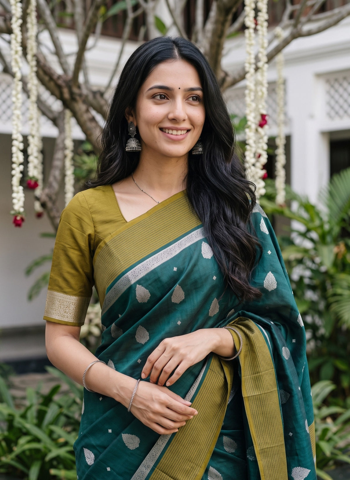 A woman wearing a dark teal green and olive yellow saree with silver motifs, standing outdoors in front of a decorated white building and hanging flower garlands.