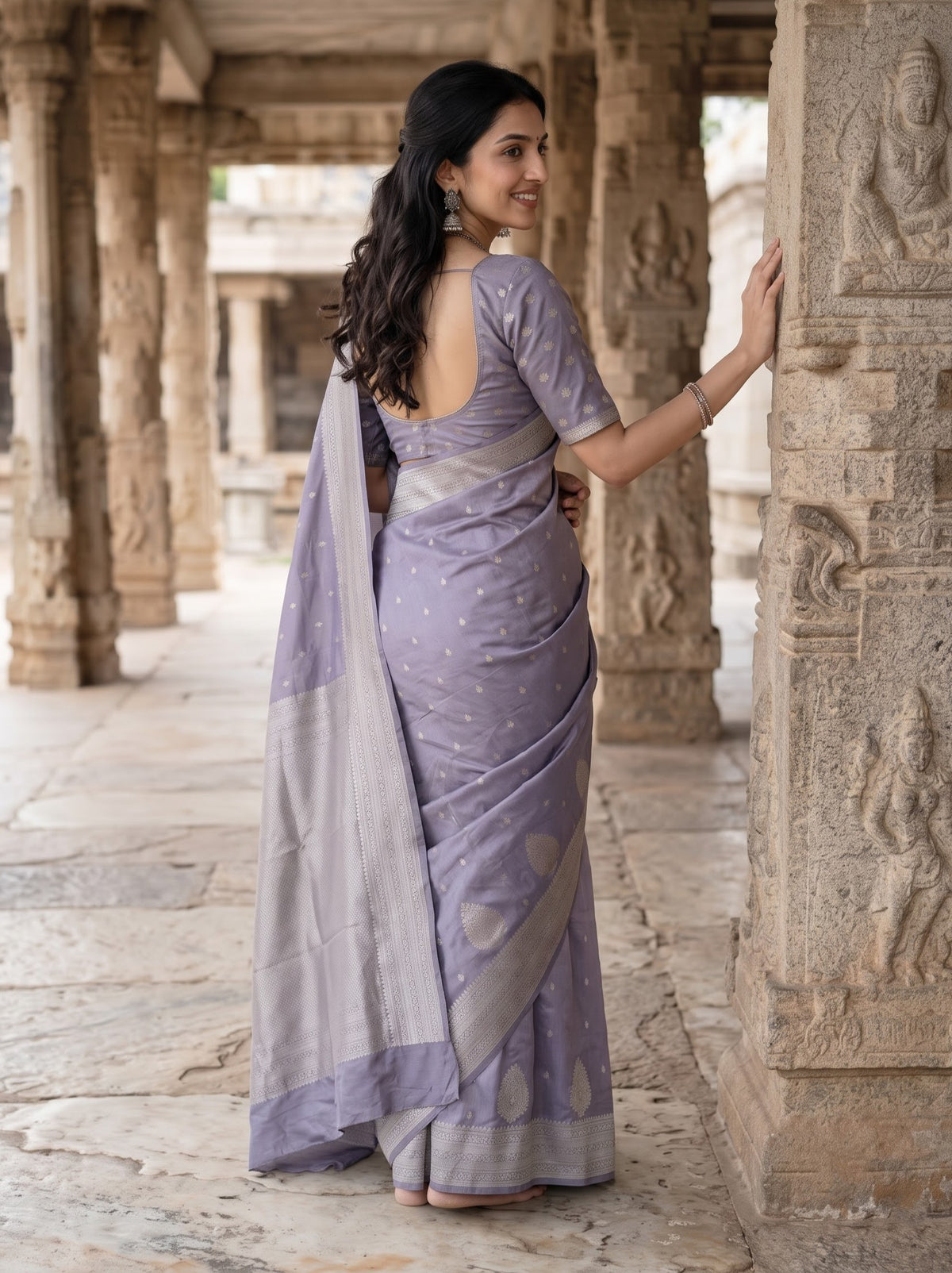 A woman wearing a lavender purple saree with a matching blouse, standing in a stone corridor with carved pillars.